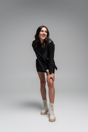 full length of happy young woman in stylish black leather jacket and skirt posing on greyの写真素材