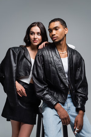 young woman in leather outfit and stylish african american man posing near high chair isolated on greyの写真素材