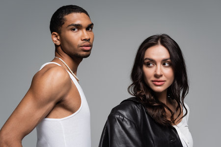 young woman in leather jacket looking at african american man in tank top isolated on grey