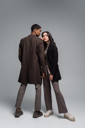 full length of stylish interracial couple in autumnal outfits posing together on greyの写真素材