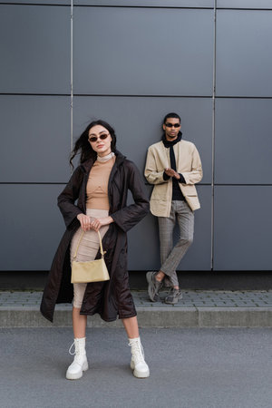 full length of stylish young woman in autumnal coat and sunglasses standing with handbag near african american manの写真素材