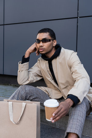 african american man in sunglasses and autumnal outfit sitting with coffee to go near shopping bagの写真素材