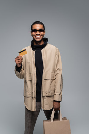 happy african american man in stylish sunglasses and autumnal outfit holding credit card isolated on greyの写真素材