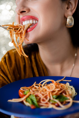 partial view of woman with red lips and golden earring eating pasta from blue plate on shiny backgroundの写真素材