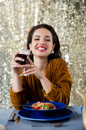 joyful and elegant woman with glass of red wine looking at camera near spaghetti on glitter silver backgroundの写真素材