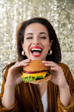 laughing woman in golden earrings holding delicious burger on shiny silver backgroundの写真素材