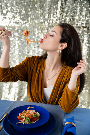 brunette woman eating tasty spaghetti near blue plates on shiny silver backgroundの写真素材
