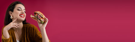 joyful and fashionable woman holding tasty burger isolated on red, banner