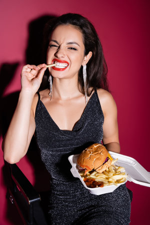 cheerful woman in black elegant dress eating french fries near burger in plastic container on red backgroundの写真素材