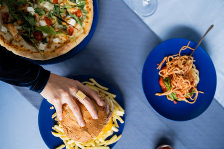 top view of cropped woman taking burger near french fries and plates with pasta and pizza on blue...