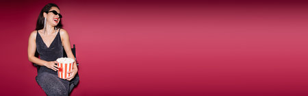 stylish woman in lurex dress and black sunglasses sitting with bucket of popcorn and laughing on red background, bannerの写真素材