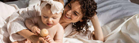 happy woman lying on bed near infant daughter in headband with wooden toy, bannerの写真素材