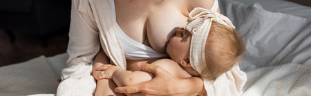 mother in loungewear breastfeeding infant daughter in headband with bow, bannerの写真素材