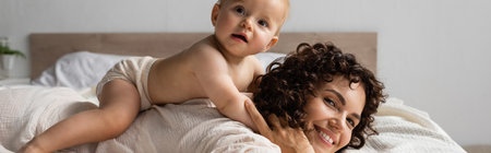 infant child lying on back of happy mother with curly hair in bedroom, banner