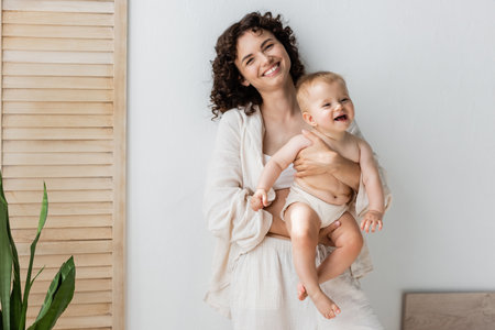 Brunette woman smiling at camera while holding baby girl near folding screen at home