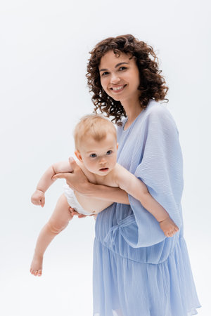 Curly woman in blue dress holding infant daughter in panties isolated on whiteの写真素材