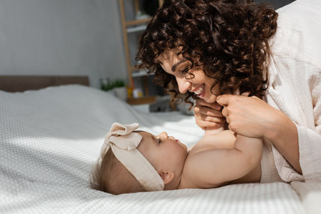 side view of cheerful mother with curly hair holding hands of baby girl in headbandの写真素材