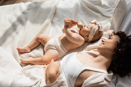 top view of happy mother lying on bed and looking at baby girl in headband with bow holding wooden rattleの写真素材