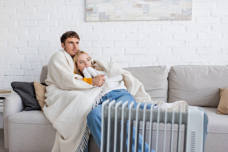 young couple covered in warm blanket hugging on couch near radiator heater at home