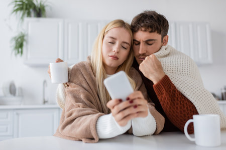 blonde young woman using mobile phone near boyfriend and cup on tableの写真素材