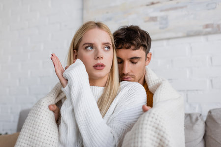 caring man covering blonde girlfriend in white sweater with blanket at homeの写真素材