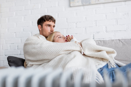 young couple covered in warm blanket hugging on couch near blurred radiator heater at homeの写真素材