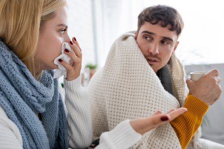 sick blonde woman in scarf sneezing in tissue near diseased boyfriend with cup of teaの写真素材