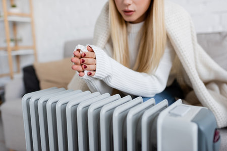 cropped view of young blonde woman covered in blanket warming hands near radiator heaterの写真素材