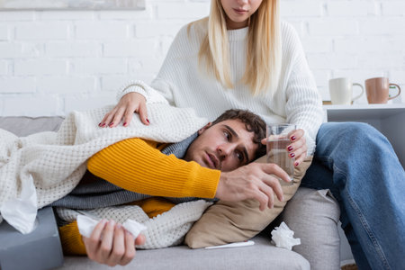 blonde woman sitting on sofa and giving glass of water to sick boyfriend