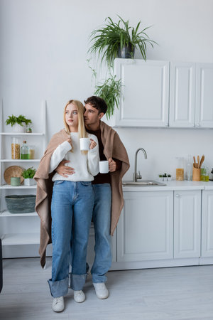 young couple covered in blanket hugging and holding cups of teaの写真素材