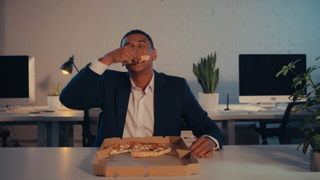 African american manager in suit eating takeaway pizza near box in office in eveningの写真素材