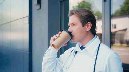 doctor in white coat drinking coffee near building of city clinic