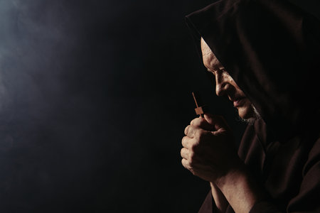 side view of monk in hood praying with holy crucifix on black backgroundの写真素材
