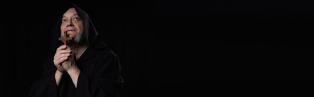 senior monk in cassock praying with wooden cross and looking up isolated on black, bannerの写真素材