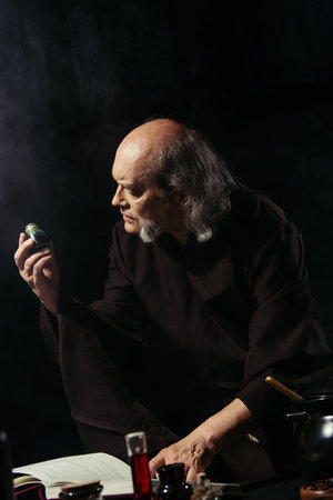 senior alchemist holding vial with magic ingredient near flasks and magic cookbook isolated on black