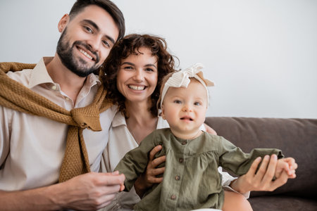joyful parents and infant girl looking at camera while sitting on sofa in living room