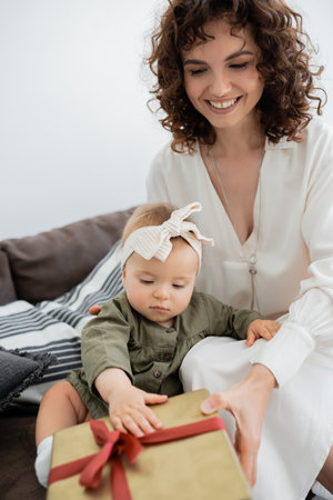 joyful mother holding wrapped gift box near infant daughter in headband sitting on sofaの写真素材