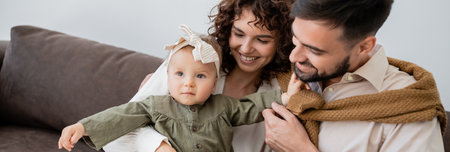 positive parents looking at infant girl in headband in living room, banner