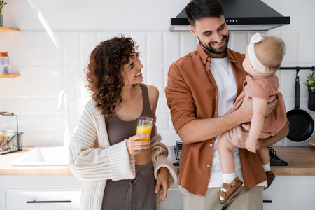 pleased man holding baby daughter near curly wife with glass of fresh orange juice in kitchenの写真素材