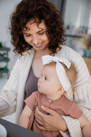 positive mother with curly hair smiling while holding infant girl in headbandの写真素材