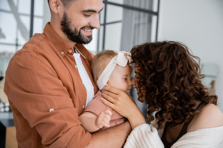 curly mother touching nose of baby daughter in headband near cheerful husband at homeの写真素材