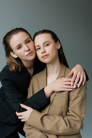 young and pretty lesbian women in black and beige blazers looking at camera isolated on grey