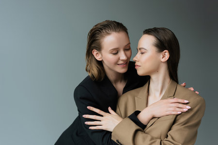 smiling lesbians in beige and black blazers hugging isolated on grey
