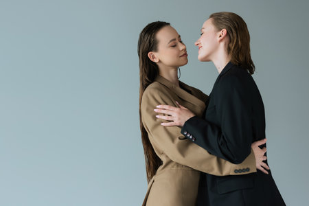 young and happy lesbian women in blazers embracing face to face isolated on greyの写真素材