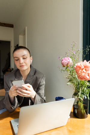 Young woman in earphone using smartphone near laptop and bouquet on table at home