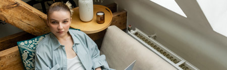 High angle view of young woman looking at camera near laptop at home, banner