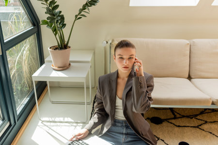 Young woman in blazer talking on smartphone and holding laptop in living roomの写真素材