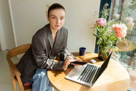 Woman in jacket looking at camera while using gadgets near flowers at homeの写真素材
