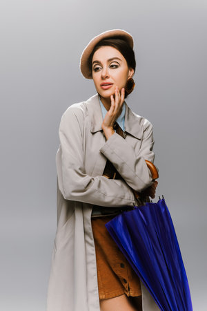 brunette young woman in beret and stylish trench coat posing with blue umbrella isolated on greyの写真素材