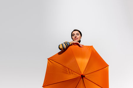 dreamy young woman posing with bright orange umbrella isolated on greyの写真素材